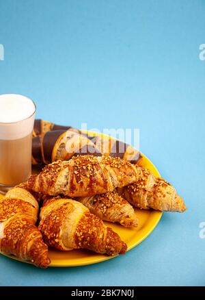 Croissants auf einem gelben Kalbsleder. Blauer Hintergrund mit Platz für Text. Ein großes Glas mit einem heißen Morgengetränk - Latte Kaffee mit Milch. Appetitlich Stockfoto