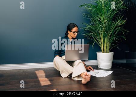 Junge Unternehmerin arbeitet auf Laptop, während sie auf dem Boden zu Hause sitzen. Geschäftsfrau, die Dokumente anschaut, während sie im Wohnzimmer an der Wand sitzt. Stockfoto
