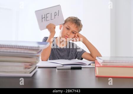 Verärgert müde preteen Kind sitzt am Tisch, seine Hausaufgaben unter Haufen von Büchern. Word Help ist auf offenem Notizbuch geschrieben Stockfoto