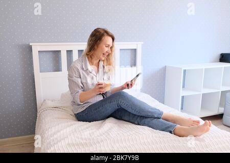 Junge Frau trinkt Kaffee und spricht auf dem Smartphone, während sie auf dem Bett sitzt Stockfoto