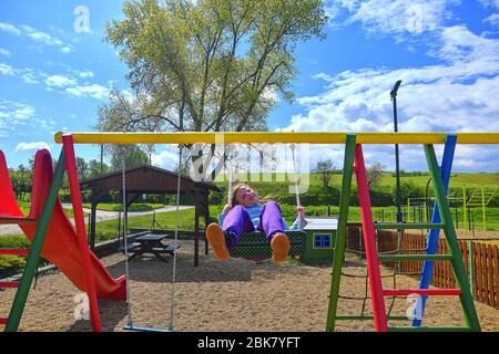 Kinder spielen auf dem Spielplatz im Freien. Kinder spielen auf der Schule oder Kindergarten Hof. Aktives Kind auf farbenfroher Schaukel. Gesunde Sommeraktivität für Kinder in Stockfoto