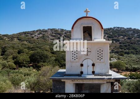 Typischer Schrein, der die Straßen in Griechenland säumen. Miniatur einer weißen Kirche mit Kreuz. Berg mit Olivenbäumen im Hintergrund. Strahlend blauer Himmel. Cr Stockfoto