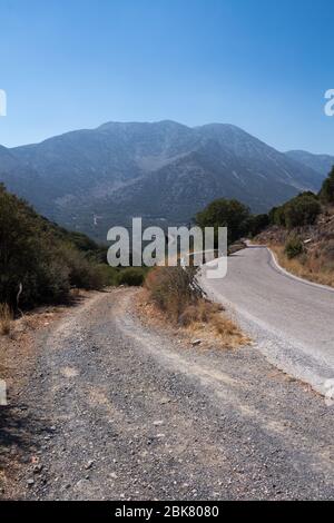 Land von Zentral-Kreta, mit einem hohen Berg im Hintergrund. Linie und Kurve eine Straße, von Steinboden, Bäumen und Büschen gesäumt. Strahlend blauer Himmel. Kreta Stockfoto