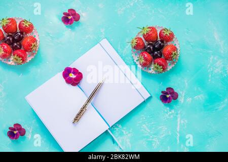 Draufsicht der offenen leeren Notizbuch, Mock up über blauen Schreibtisch Hintergrund. Waldbeeren Obst Arrangement.Erdbeeren, Konzept der geschäftigen Frühstück am Morgen Stockfoto