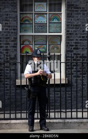 Ein Polizeibeamter steht vor "Regenbogenzeichnungen" in einem Fenster der Downing Street Nr. 10 in London, um NHS-Arbeiter gegen das Coronavirus in Großbritannien zu unterstützen Stockfoto