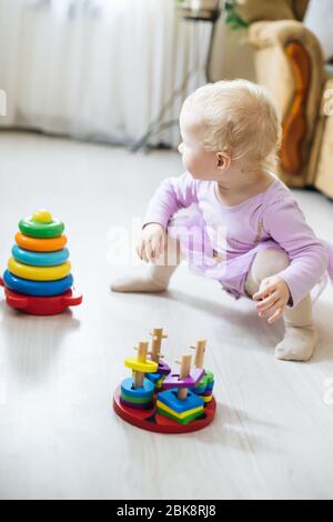 Mädchen spielt logische Pyramide auf dem Boden im Wohnzimmer am sonnigen Tag. Montessori Holzspielzeug gefaltete Pyramide. Kreis, Quadra, Dreieck, rechteckiges Holzelem Stockfoto