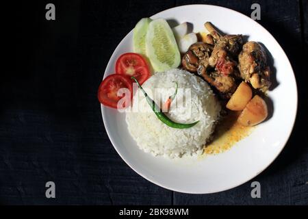 Tasse gekochter Reis mit Hühnerbrühe auf einem weißen Teller auf schwarzem Hintergrund isoliert. Gekochter Reis auf einem weißen Teller, Chili, Tomaten, Gurke. Stockfoto