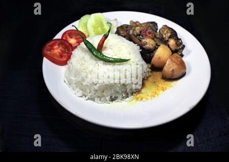 Tasse gekochter Reis mit Hühnerbrühe auf einem weißen Teller auf schwarzem Hintergrund isoliert. Gekochter Reis auf einem weißen Teller, Chili, Tomaten, Gurke. Stockfoto