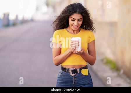 Arabisches Mädchen, das mit einem Kaffee zum Mitnehmen auf der anderen Straßenseite spazieren geht Stockfoto