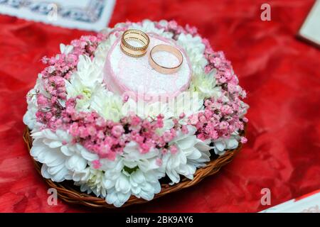 Zwei goldene Eheringe auf einem schönen weißen und rosa Bouquet in rustikalem Korbflecht. Roter Stoffhintergrund. Nahaufnahme. Stockfoto