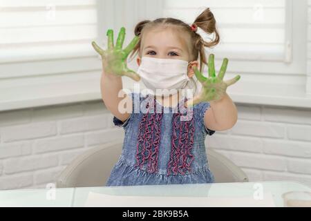 Kleines Mädchen in Maske malt mit Fingern.Mädchen lächelt und hob ihre grünen Palmen in der Farbe, Selbstisolierung, Coronavirus covid-19, zu Hause bleiben Stockfoto