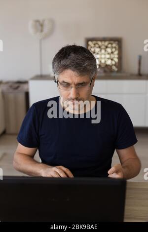 Vertikales Porträt eines Mannes mittleren Alters mit Brille, der von zu Hause aus mit schwarzem Laptop arbeitet. Stockfoto