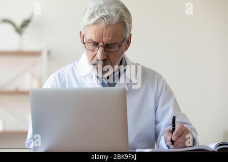 Seriöse alte Arzt Schreiben Notizen, mit Laptop in der medizinischen Praxis Stockfoto