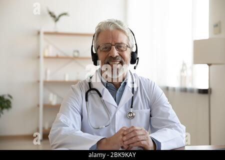 Lächelnder Oberarzt mit Headset, der sich auf Kamera und Telemedizin-Konzept besieht Stockfoto