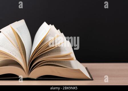 Ein großes, dickes Hardcover-Buch liegt auf einem hellen Holztisch. Das Buch ist offen, die Blätter der Seite sind ausgespannt, und die Rückseite ist ein schwarzer Hintergrund Stockfoto