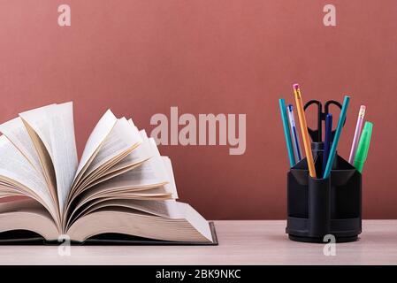 Ein Buch und ein Ständer mit Stiften, Bleistiften und Scheren auf einem hellen Holztisch. Das Buch ist offen, die Blätter der Seite sind ausgespannt, ein braunes schwarzes Backg Stockfoto