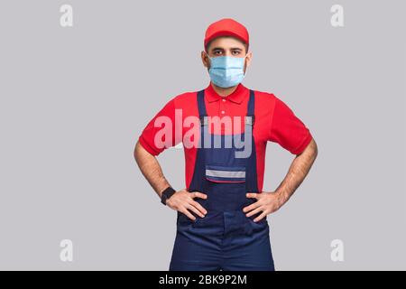 Porträt des jungen Mannes mit chirurgischer medizinischer Maske in Blau insgesamt, rotes T-Shirt und Kappe stehen und halten die Hände auf der Taille und schauen auf Kamera, indoo Stockfoto