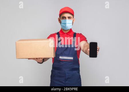 Lieferung in Quarantäne. Das ist für Sie! Junger Mann mit chirurgischer medizinischer Maske in blauer Uniform und rotem T-Shirt stehend, Karton und Schau halten Stockfoto