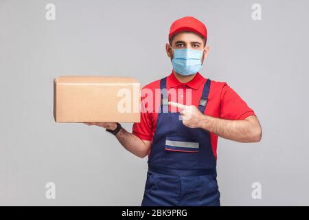 Lieferung in Quarantäne. Junger Mann mit chirurgischer medizinischer Maske in blauer Uniform und rotem T-Shirt stehend, mit dem Finger auf den Pappkarton halten und zeigen Stockfoto