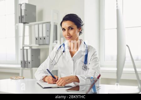 Selbstbewusste Frau Ärztin Kinderärztin schreibt in einer Zwischenablage, die an einem Tisch in einem weißen Büro des Krankenhauses sitzt Stockfoto