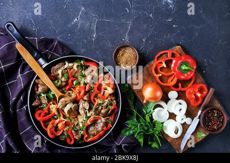 Hühnerleber mit Zwiebeln und Paprika, frische Petersilie auf der Oberseite, auf einer Pfanne mit Zutaten auf dunklem Betongrund, Draufsicht, Fla Stockfoto