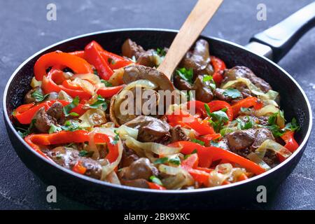 Gebratene Hühnerleber mit Zwiebelringen und roter Paprika, frische Petersilie auf einer Pfanne mit einem Holzlöffel auf dunklem Betongrund, Horizont Stockfoto