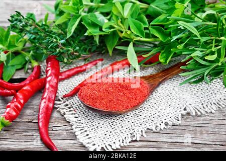 Gemahlener scharfer roter Pfeffer in einem Löffel auf Burlap, Chilischoten und würzigen Grüns auf Holzbrett Hintergrund Stockfoto