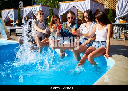 Eine Gruppe von Freunden, die am Pool Spaß haben, und die Gläser mit frischen Cocktails, die am Pool am sonnigen Sommertag Wasser spritzen, zusammenstoßen. Menschen Stockfoto