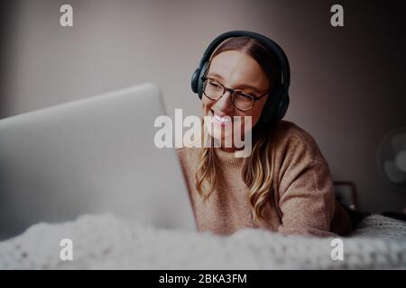 Porträt einer lächelnden jungen Frau, die Kopfhörer trägt und zu Hause Musik auf ihrem Laptop hört, während sie online auf dem Bett liegt Stockfoto