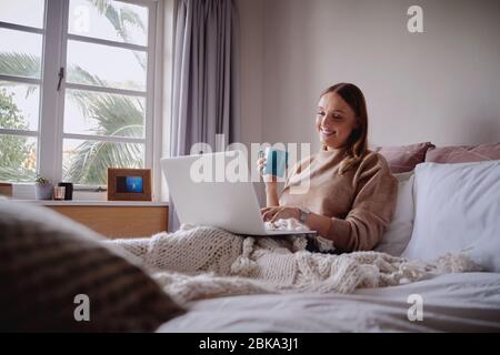 Lächelnde junge Frau genießen Morgenkaffee mit Laptop sitzen in ihrem Bett arbeiten von zu Hause aus - Student Online-Vorlesungen studieren Stockfoto