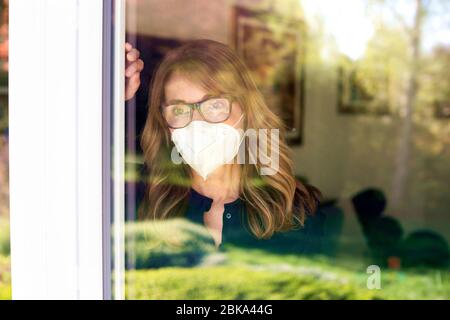 Clsoe-up-Porträt einer traurigen Frau, die am Fenster steht und eine Gesichtsmaske trägt, um sich vor einer Coronavirus-Pandemie zu schützen. Stockfoto