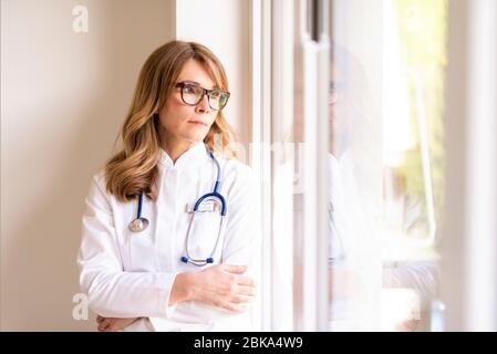 Porträt der Ärztin, die beim Fenster und beim Blick nach draußen aufmerksam schaut. Stockfoto