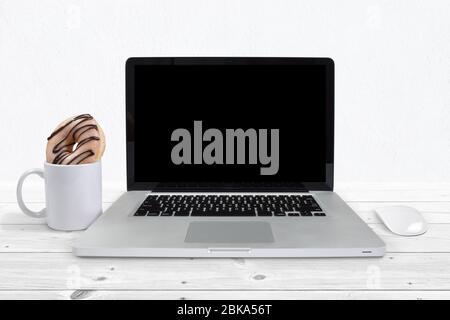 Laptop sitzt auf einer grauen Holzfläche neben einer Kaffeetasse mit einem verführerischen glasierten und Schokolade Eisdonut gefüllt. Stockfoto