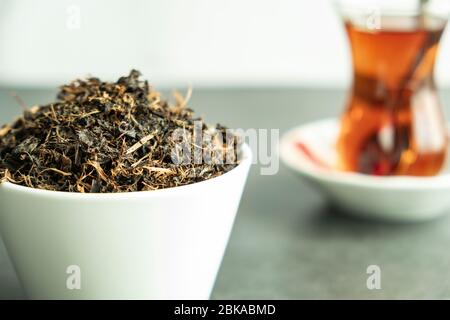 Trockenes schwarzes Teeblatt in weißer Porzellanschale und frisch gebrühter frischer Tee in traditionellem türkischen Glas auf Betonuntergrund. Traditionell, gesund Stockfoto