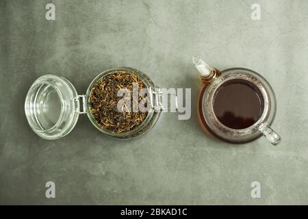 Trockenes schwarzes Teeblatt im Glasgefäß und frisch gebrühter frischer Tee in der Teekanne auf Beton, Steingrund. Traditionelles, gesundes und biologisches Getränk. Stockfoto