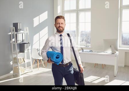 Glücklicher erfolgreicher Geschäftsmann in einem weißen Hemd mit Yogamatte im Büro. Stockfoto