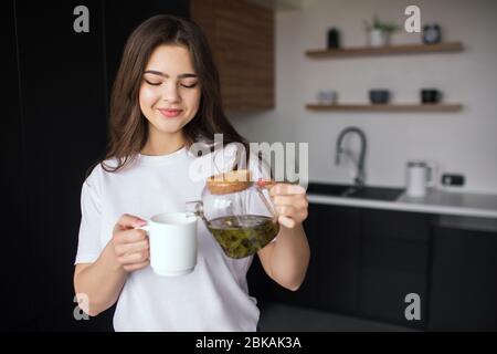 Junge Frau in der Küche während der Quarantäne. Mädchen in lässigem Look und weißes Hemd Gießen etwas Tee aus Teekanne in weiße Tasse. Ich werde es trinken. Stockfoto