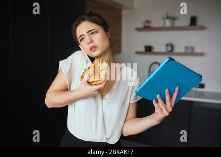 Junge Frau in der Küche während der Quarantäne. Mädchen am Telefon reden und essen Sandwich. Geschäftsfrau hält blaue Tablette in der Hand. Home Office und Remote Stockfoto