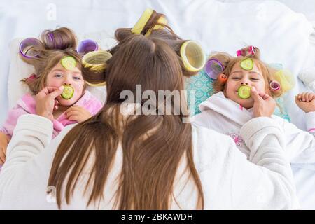 Glückliche liebevolle Familie. Mutter und Kinder machen Haare und haben Spaß. Mutter und Tochter werden Gurken auf die Augen gelegt, die im Schlafzimmer auf dem Bett sitzen. Stockfoto