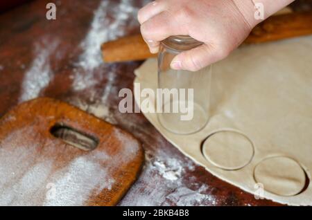 Eine weibliche Hand schneidet Kreise in einem Teigblech mit einem Glas aus. Dünn gerollter Teig für Fleischknödel auf dem Küchentisch. Der Prozess des Garens h Stockfoto