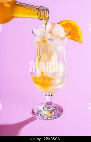 Vorbereitung eines kalten Sommergetränks durch Gießen Orangensirup über Eiswürfel in einem Glas, auf einem lila Hintergrund. Herstellung von Orangenlimonade. Tropischer Mocktail. Stockfoto