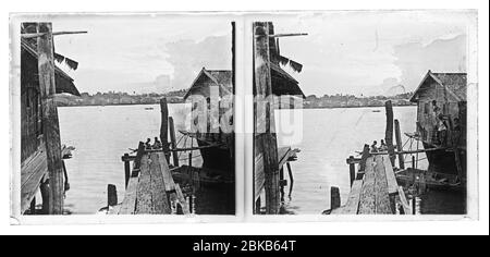 Kambodscha traditionelle Holzstelzenhäuser am Wasser am Mekong oder Tonle SAP. Kinder stehen am Hauswall und beobachten ihre Kumpel beim Paddeln in einem traditionellen Boot auf dem Fluss. Stereobild von um 1910. Bild auf trockener Glasplatte aus der Sammlung Herry W. Schaefer Stockfoto