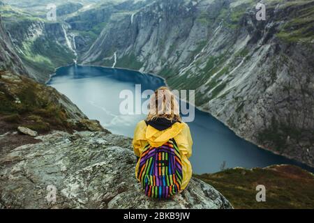 Mädchen in einer gelben Jacke sitzt am Rand einer Klippe vor der Kulisse der Berge von Norwegen und dem bewölkten Himmel Stockfoto