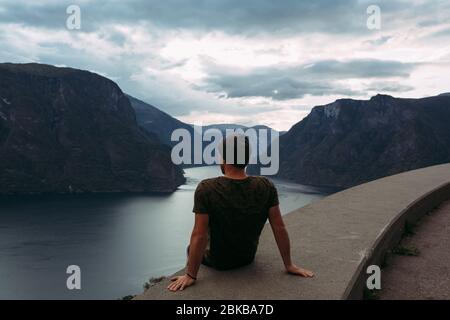 Ein Mann sitzt am Rande der Attika vor dem Hintergrund der Berge Norwegens und dem Himmel mit Wolken Stockfoto