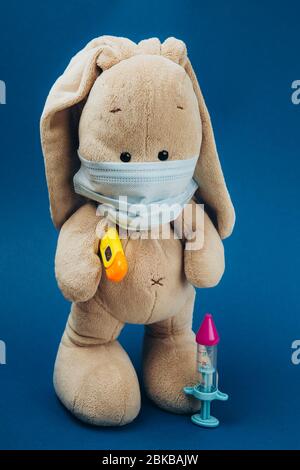 Spielzeug Hase mit Thermometer unter Pfote und in schützender medizinischer Maske, Spritze auf blauem Hintergrund. Spielzeug wurde krank .Medizin Konzept. Stockfoto