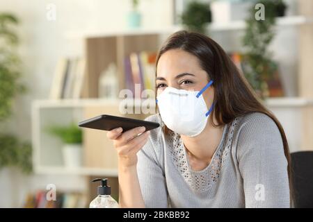 Glückliche Frau mit Spracherkennungssystem auf dem Smartphone in covid-19 Confinement trägt Maske zu Hause sitzen Stockfoto