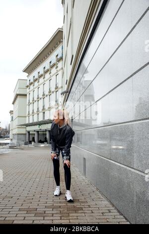 Hübsches blondes Mädchen mit Pferdeschwanz trainiert in Sportkleidung auf den Straßen einer Stadt. Sie trägt eine sportliche Jacke, Leggings und Sneaker. Stockfoto