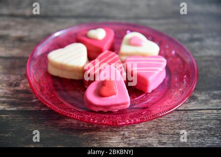 Herzförmige Pralinen in rosa, weiß und rot auf einem rosafarbenen Glasteller. Glasplatte hat ein Ornamentmuster. Dunkler Holztisch Hintergrund. Stockfoto