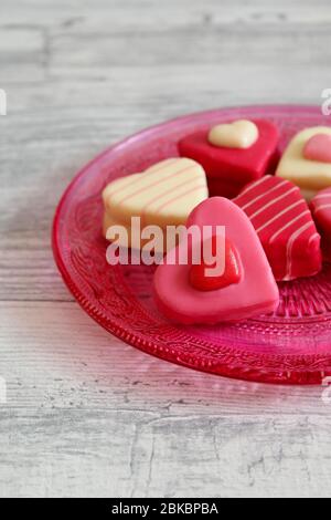 Herzförmige, bunte Pralinen (Petit Fours) auf einem rosa Glasteller mit Ornamentmuster. Heller Holztisch Hintergrund. Stockfoto