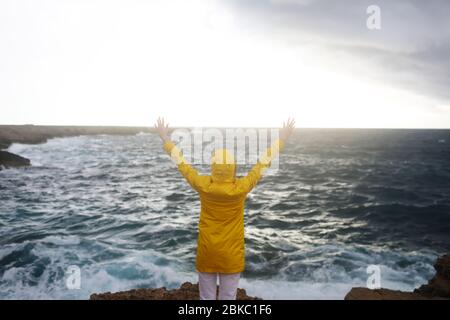 Junge Frau in gelbem Regenmantel stehend mit ausgestreckten Armen gekleidet, während Sie schöne Meereslandschaft in regnerischen Tag am Felsstrand in Wolken genießen Stockfoto